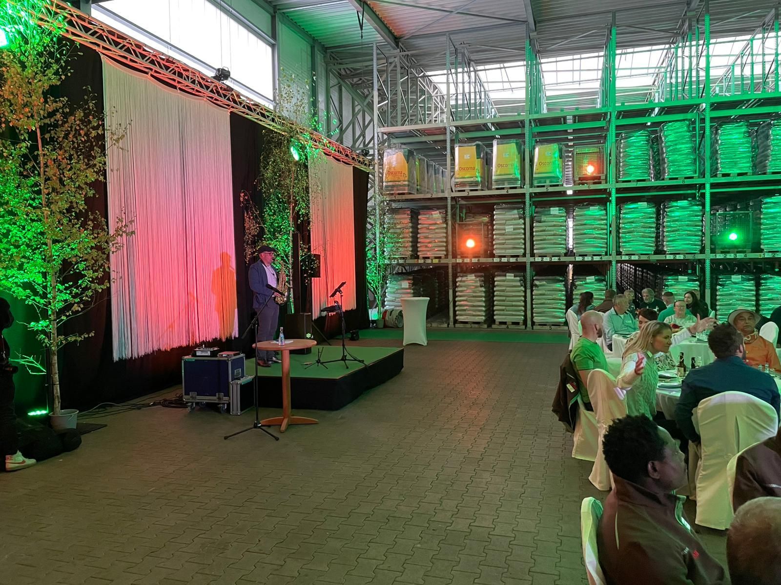 Ein Mann spricht auf der Bühne bei einer Veranstaltung in einem Lagerhaus, mit sitzendem Publikum und bunter Beleuchtung.