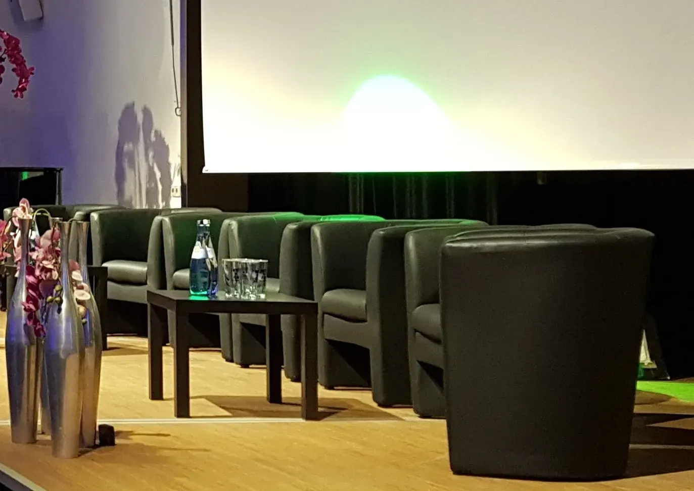 Eine Reihe von schwarzen Stühlen und Tischen auf einer Bühne vor einem Bildschirm