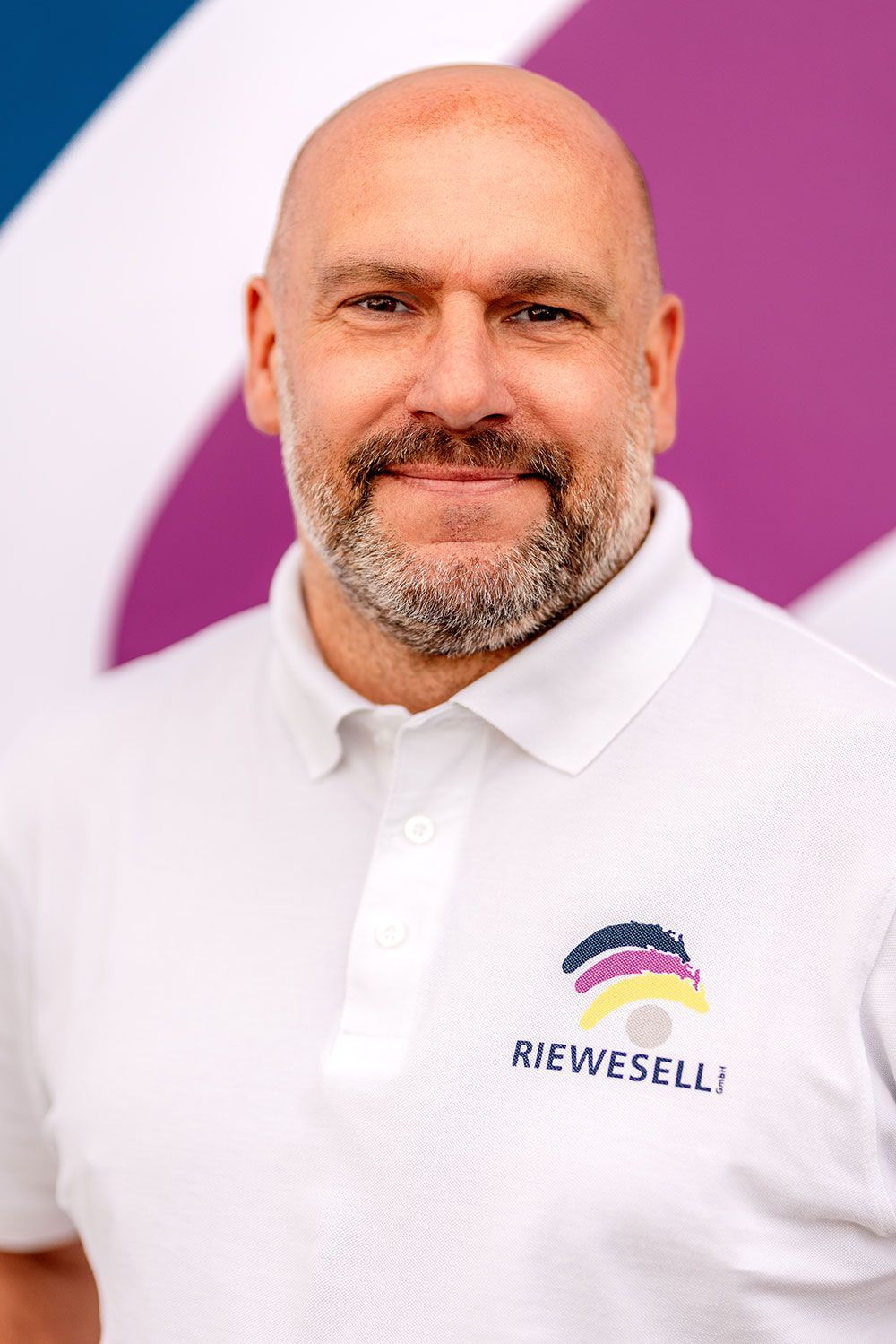 Ein Mann mit Glatze und Bart, der ein weißes Poloshirt mit Logo trägt, lächelt vor einem farbenfrohen Hintergrund.