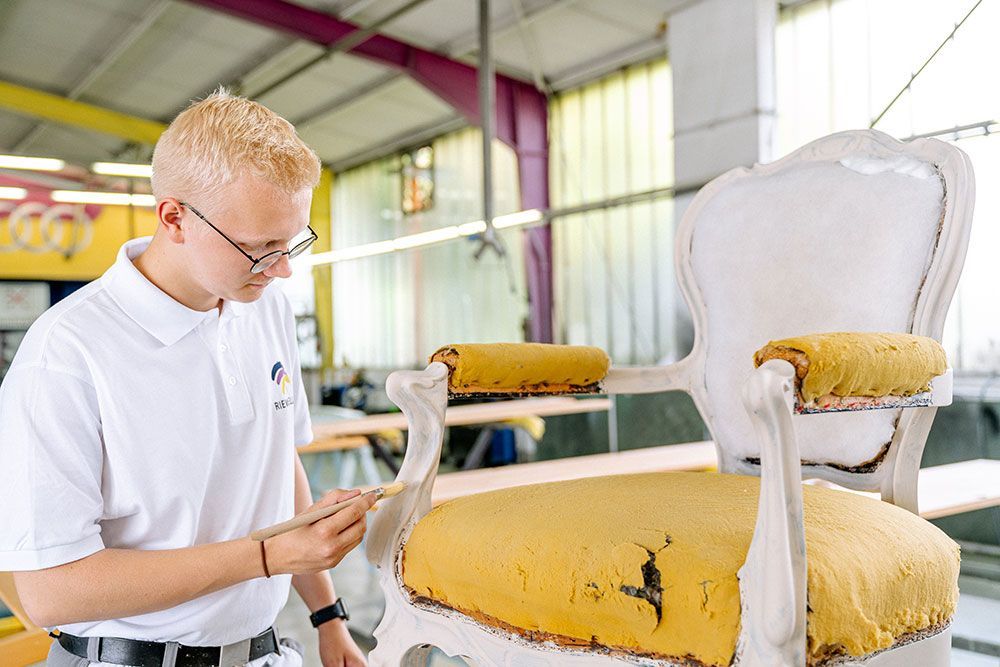 Eine Person arbeitet in einer Werkstatt an einem antiken Stuhl und bemalt dessen Gestell.