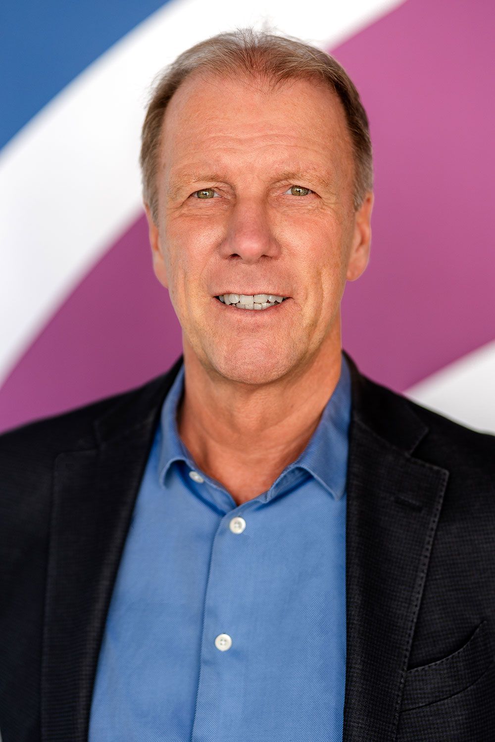 Ein Mann in blauem Hemd und schwarzer Jacke lächelt vor einem farbenfrohen, geschwungenen Hintergrund.