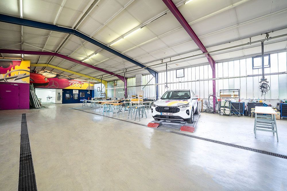 Innenansicht einer großen Werkstatt mit einem Auto auf einer Hebebühne. Werkzeuge und Ausrüstung sind verstreut.