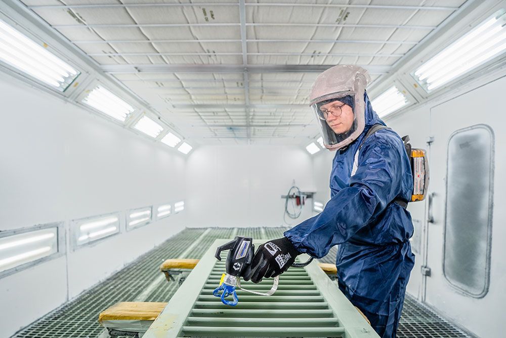 Eine Person in Schutzkleidung besprüht einen Metallrahmen in einer Spritzkabine.