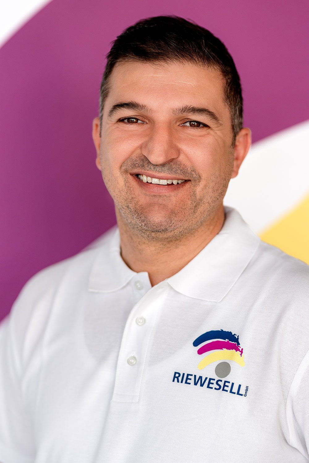 Mann im weißen Poloshirt lächelt. Bunter, abstrakter Hintergrund mit dem Logo „RIEWESELL“.