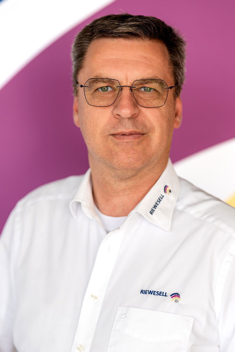 Mann mit Brille und weißem Hemd mit Logo, vor einem lila-gelben Hintergrund.
