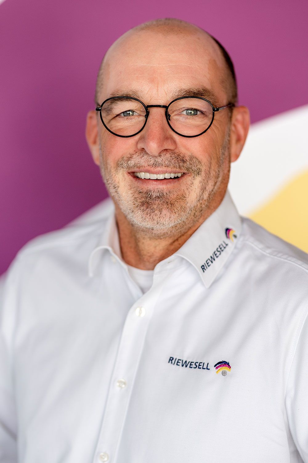 Mann mit Brille lächelt, er trägt ein weißes Hemd mit Firmenlogo. Lila-gelber Hintergrund.