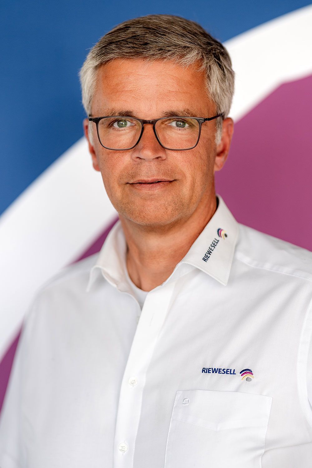Mann mit Brille und weißem Hemd mit Logo vor einem blau-violetten Hintergrund.