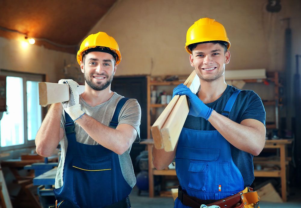 Zwei Arbeiter in gelben Schutzhelmen und blauen Overalls tragen Holz. Sie lächeln in einer Werkstatt.