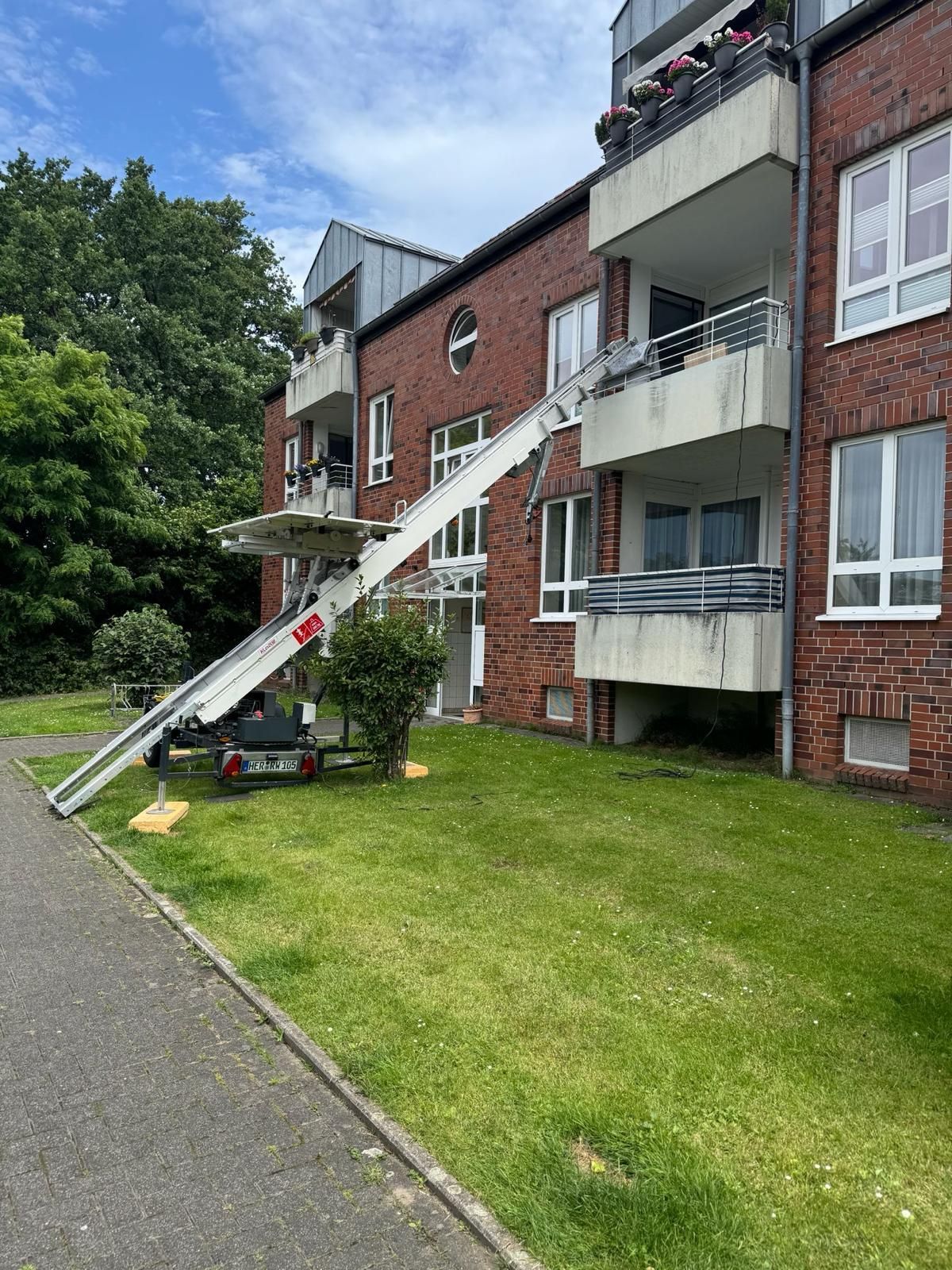 Wohnhaus mit einer bis zu einem Balkon ausgefahrenen Dachleiter auf einer Rasenfläche neben einem gepflasterten Weg.