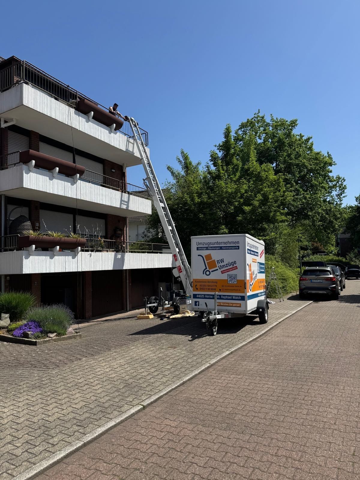 Weißer Umzugswagen, der neben einer angelehnten Leiter und Bäumen vor einem Mehrfamilienhaus geparkt ist