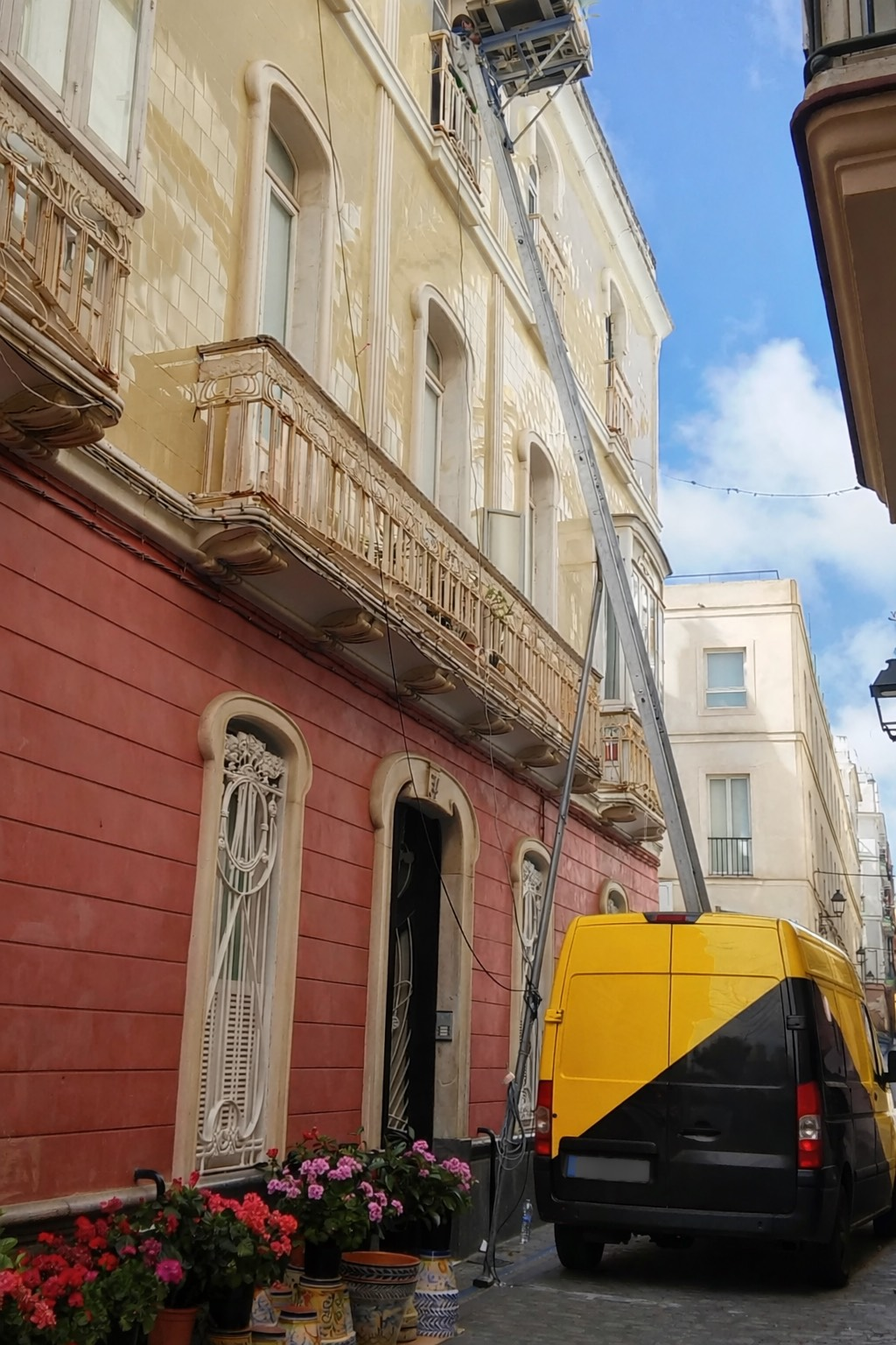 Furgoneta amarilla y negra con elevador que ayuda a trasladar mercancías desde un edificio histórico con balcones.
