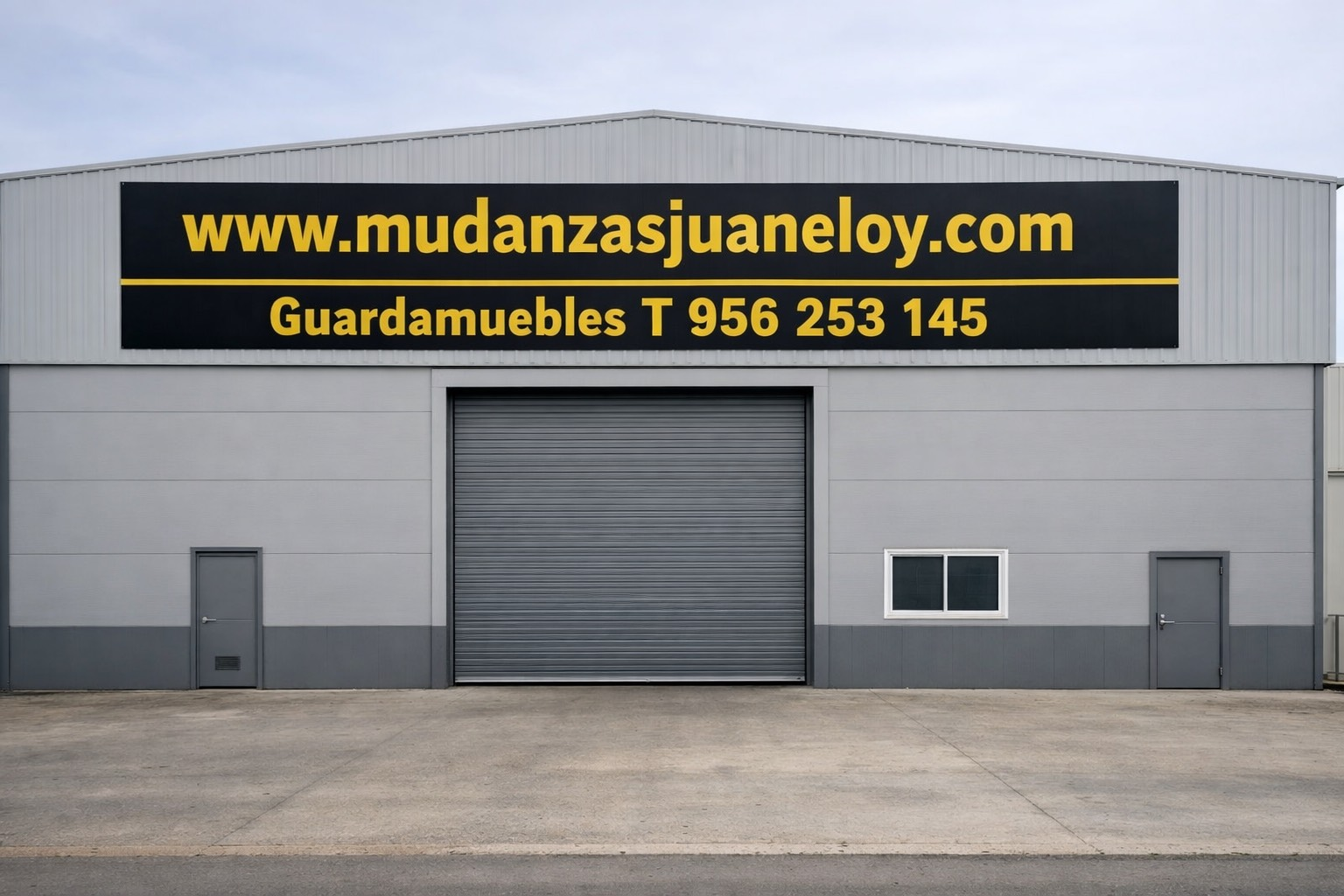 Almacén gris con una puerta grande, dos puertas más pequeñas, una ventana y un cartel: www.mudanzasjuaneloy.com