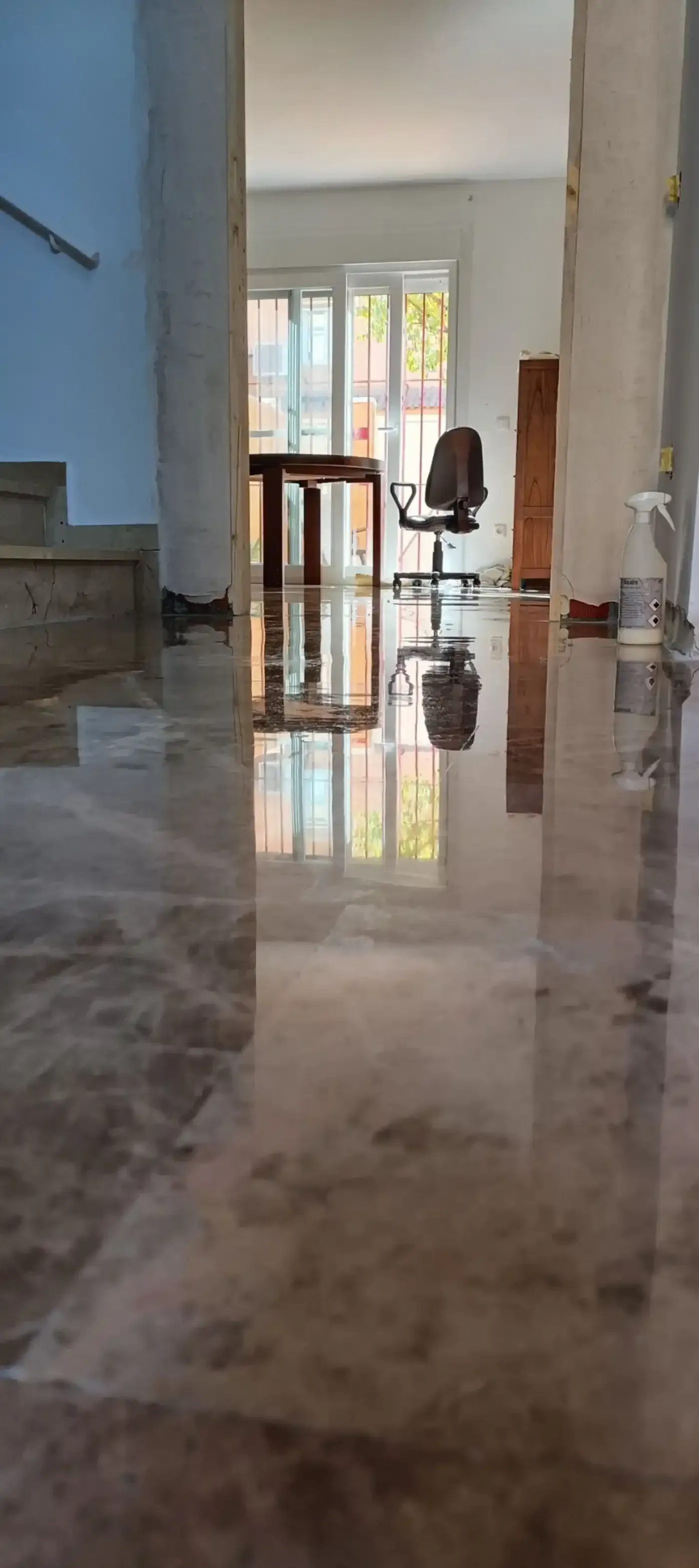 Piso reflectante en una habitación, que refleja una mesa, una silla y la luz del sol a través de una ventana.
