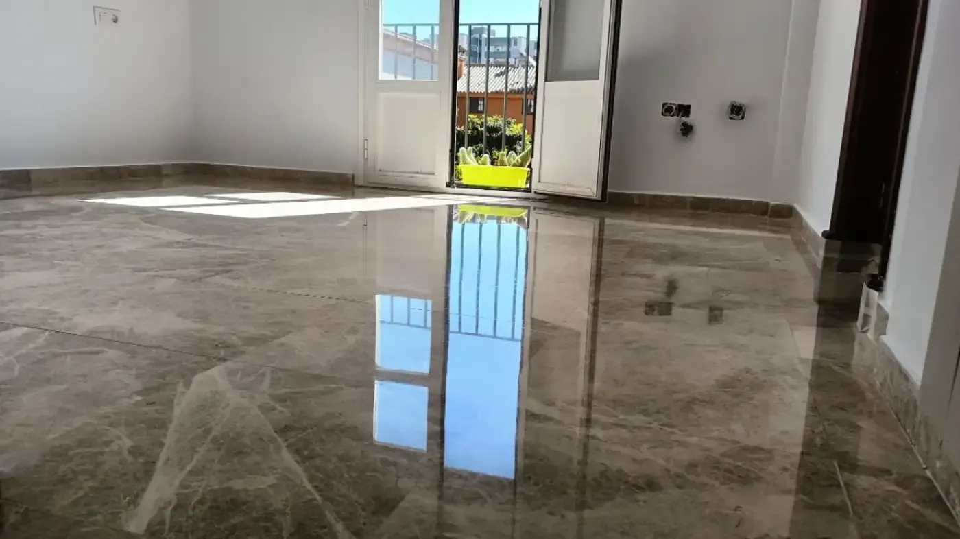 Habitación vacía con suelo marrón brillante, paredes blancas y puerta abierta al exterior.