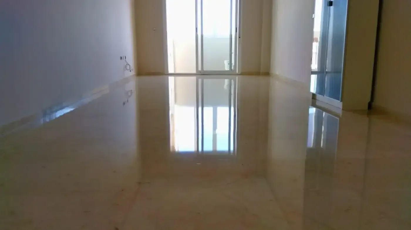 Habitación vacía con piso de baldosas beige brillante que refleja la luz del sol desde una puerta de vidrio.