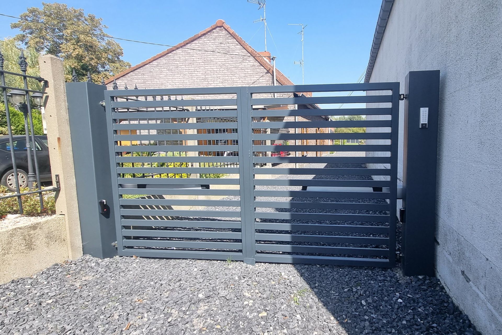Un portail métallique gris se dresse devant une allée de gravier menant à un bâtiment.