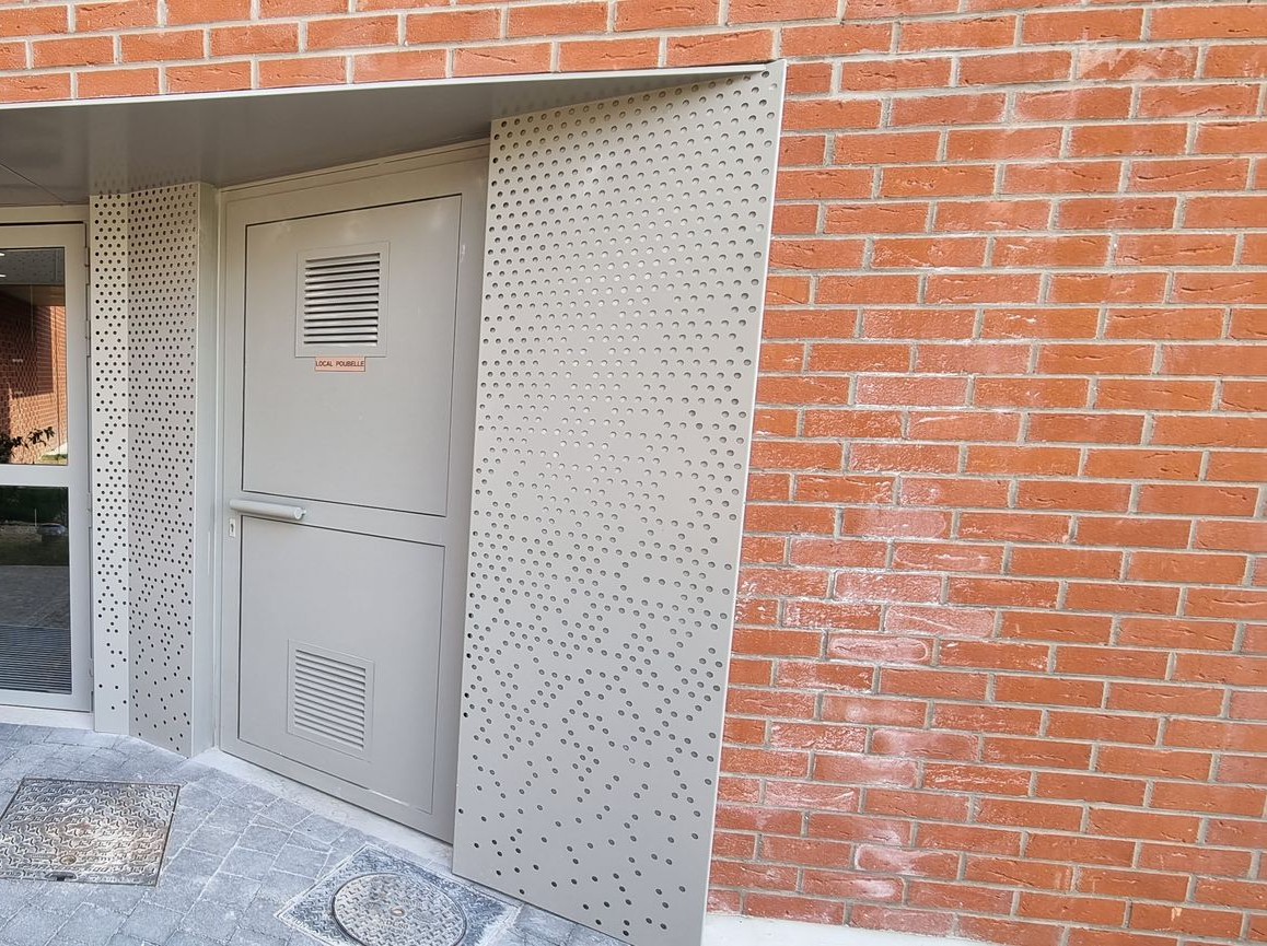 Une porte en métal beige à panneaux perforés, encastrée dans l'entrée d'un bâtiment en briques rouges.