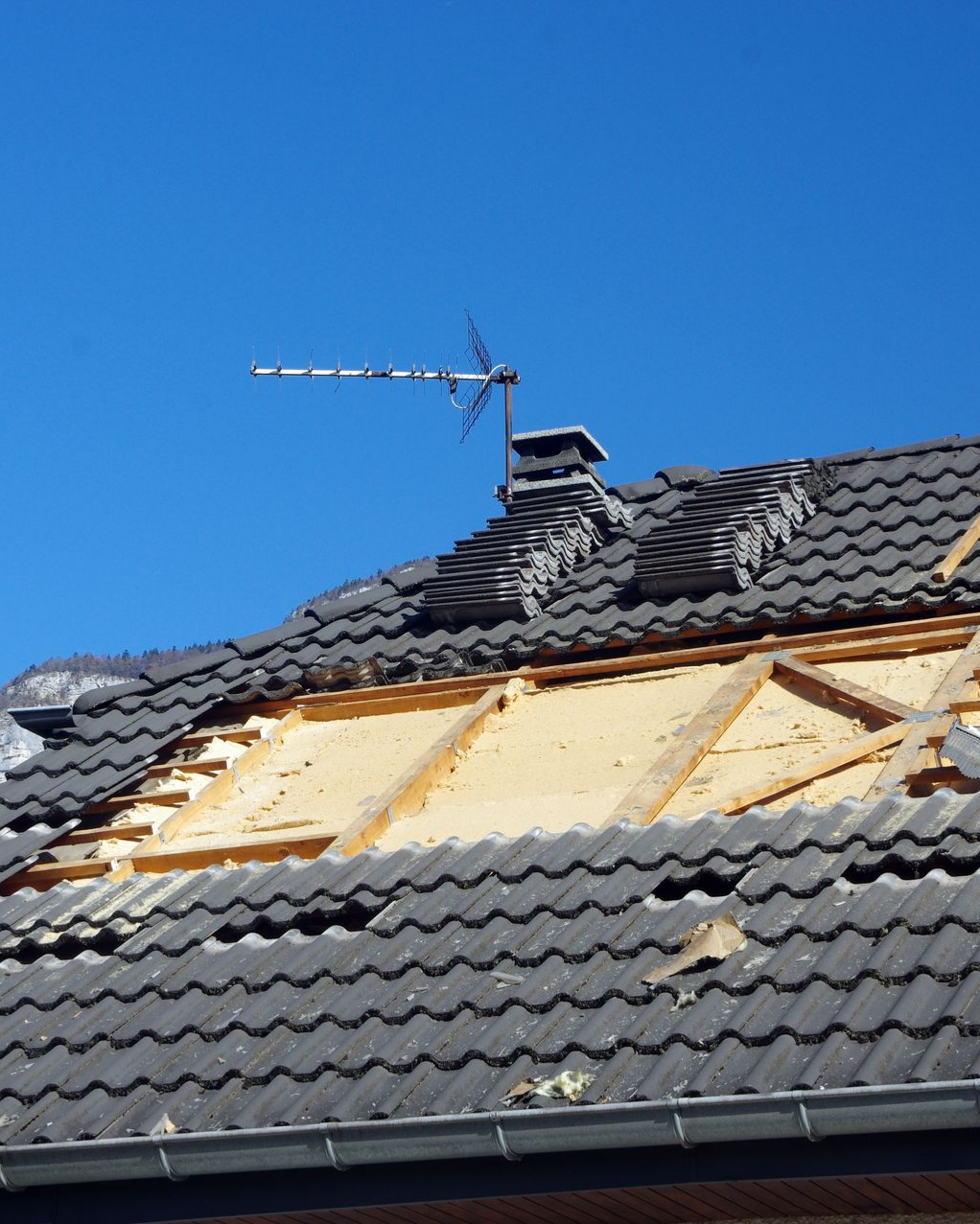 Toiture partiellement arrachée, laissant apparaître les poutres en bois et l'isolant. Une antenne de télévision se dresse sur les tuiles restantes, sur fond de ciel bleu.