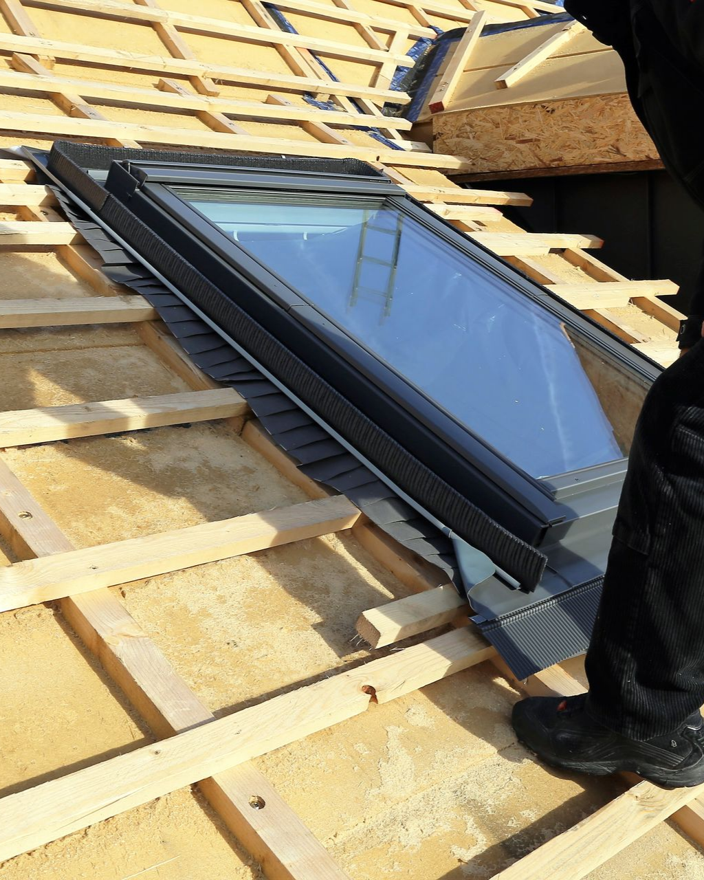 Installation d'un puits de lumière sur une charpente de toit en bois. Une personne en pantalon et chaussures sombres travaille sur la charpente.