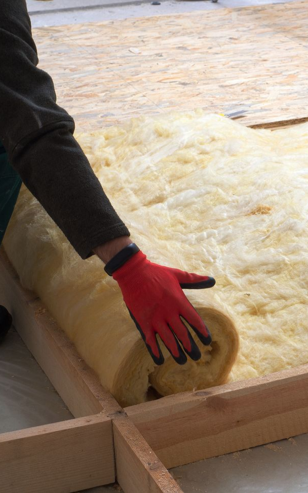 Une personne portant des gants rouges déroule de l'isolant sur un plancher à ossature bois.