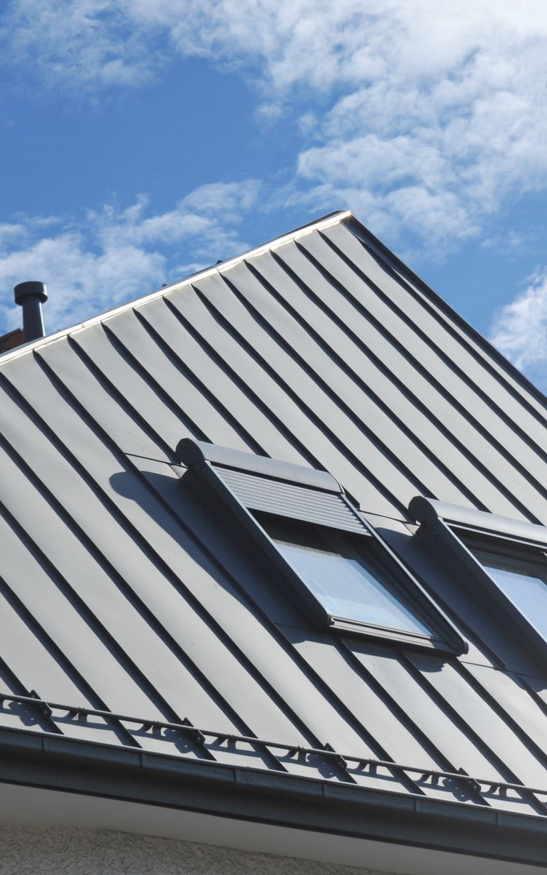 Toit en métal gris avec puits de lumière, sous un ciel bleu nuageux.