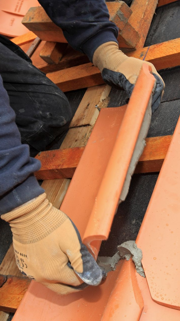 Une personne portant des gants pose une tuile rouge sur un toit avec du mortier.