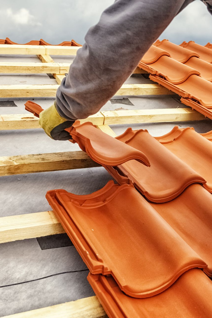 Un couvreur pose des tuiles de terre cuite rouge sur une charpente en bois.