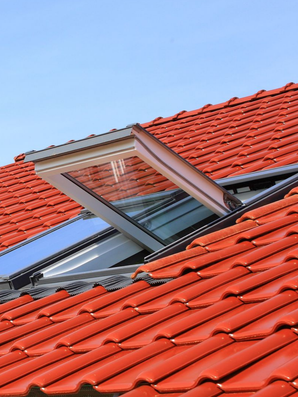 Toit de tuiles rouges avec une lucarne ouverte sur un ciel bleu.