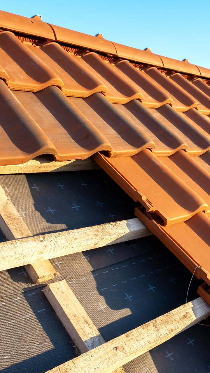 Gros plan sur une toiture en construction : tuiles en terre cuite rouge posées sur une sous-couche noire et des poutres de soutien en bois.