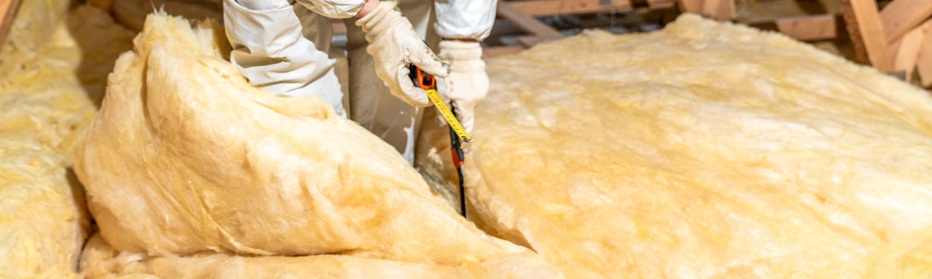 Une personne portant un équipement de protection découpe l'isolant dans un grenier. On aperçoit de l'isolant beige et des poutres en bois.