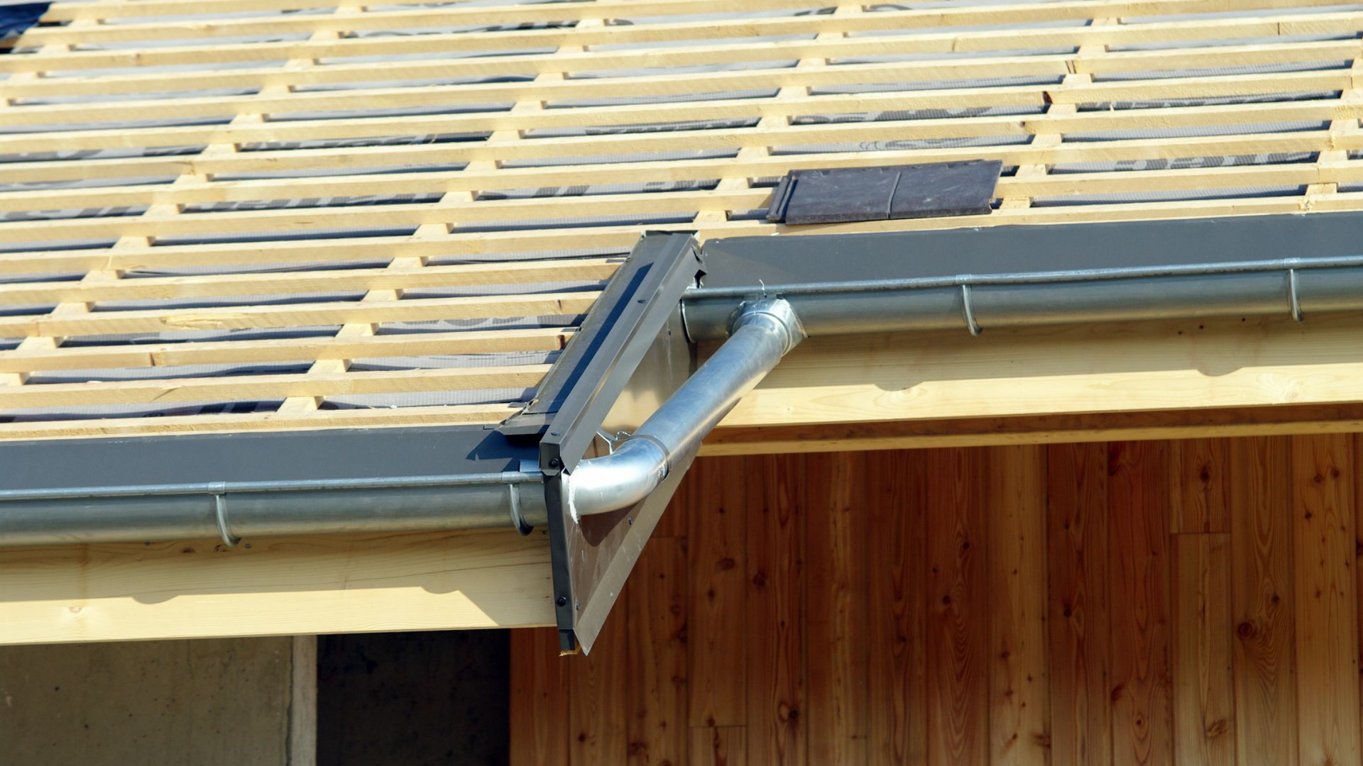 Toiture en construction avec poutres en bois, gouttières grises et descente pluviale contre un mur en bois.