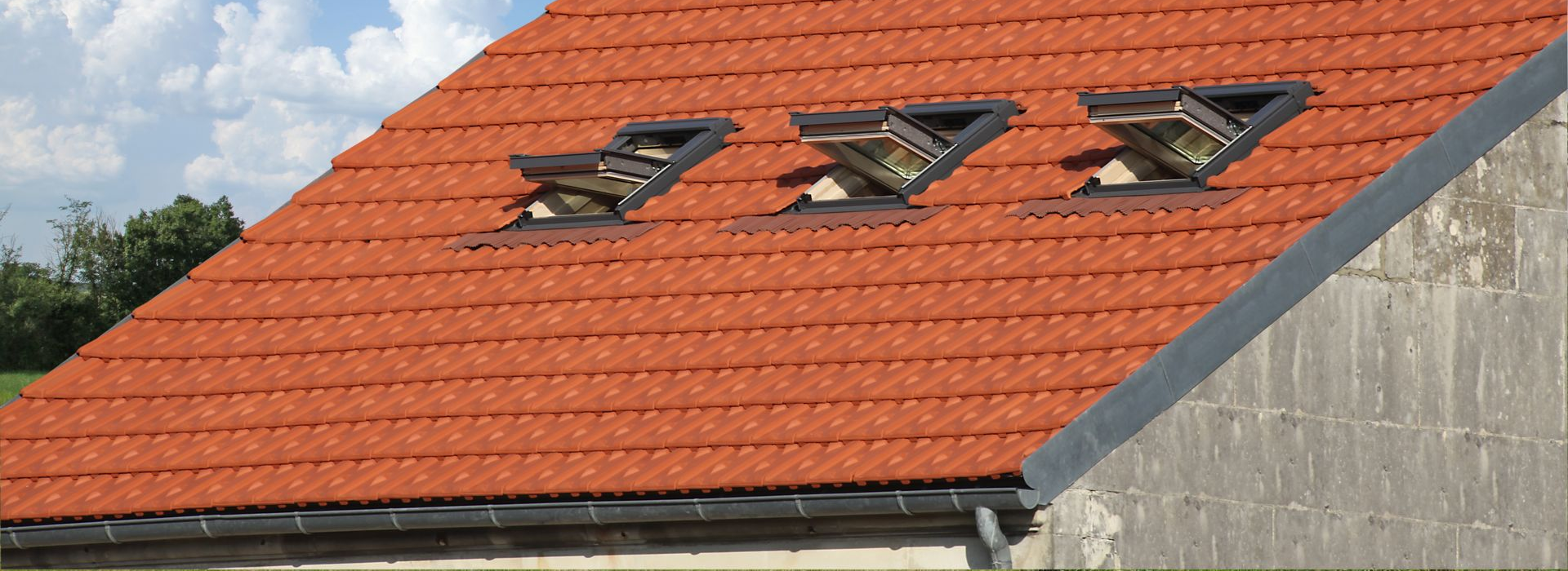 Toit de tuiles rouges percé de trois puits de lumière sur un bâtiment dont le ciel est nuageux en arrière-plan.