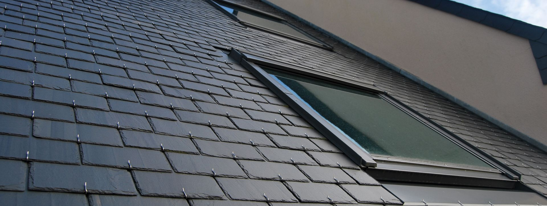 Toit en ardoise noire avec une lucarne et un bâtiment clair en arrière-plan.