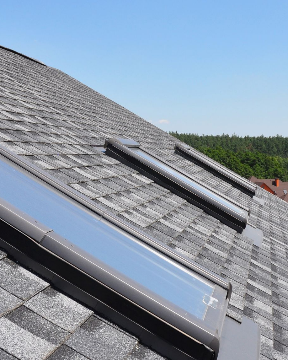 Trois puits de lumière sur un toit en bardeaux d'asphalte gris se détachant sur un ciel bleu, avec une ligne d'arbres visible au loin.