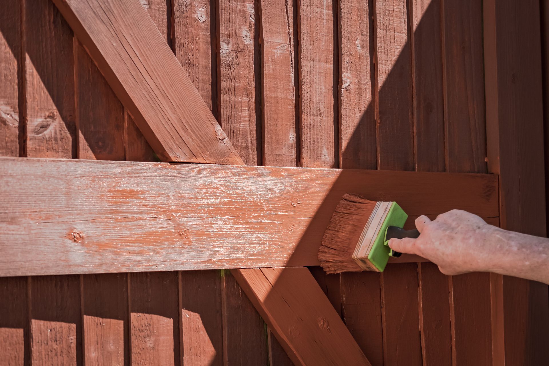 Une personne utilise un pinceau pour peindre une clôture en bois brun, faisant apparaître un contraste net entre le vieux bois patiné et la peinture neuve.