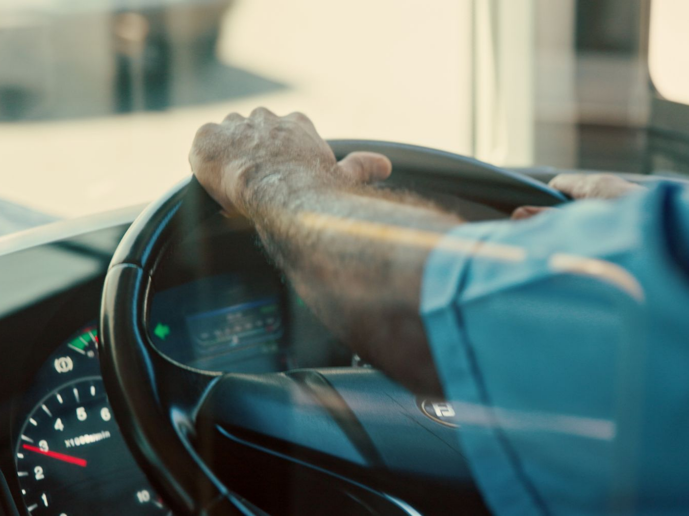 Un conducteur de bus en chemisette bleue a les mains sur le volant.