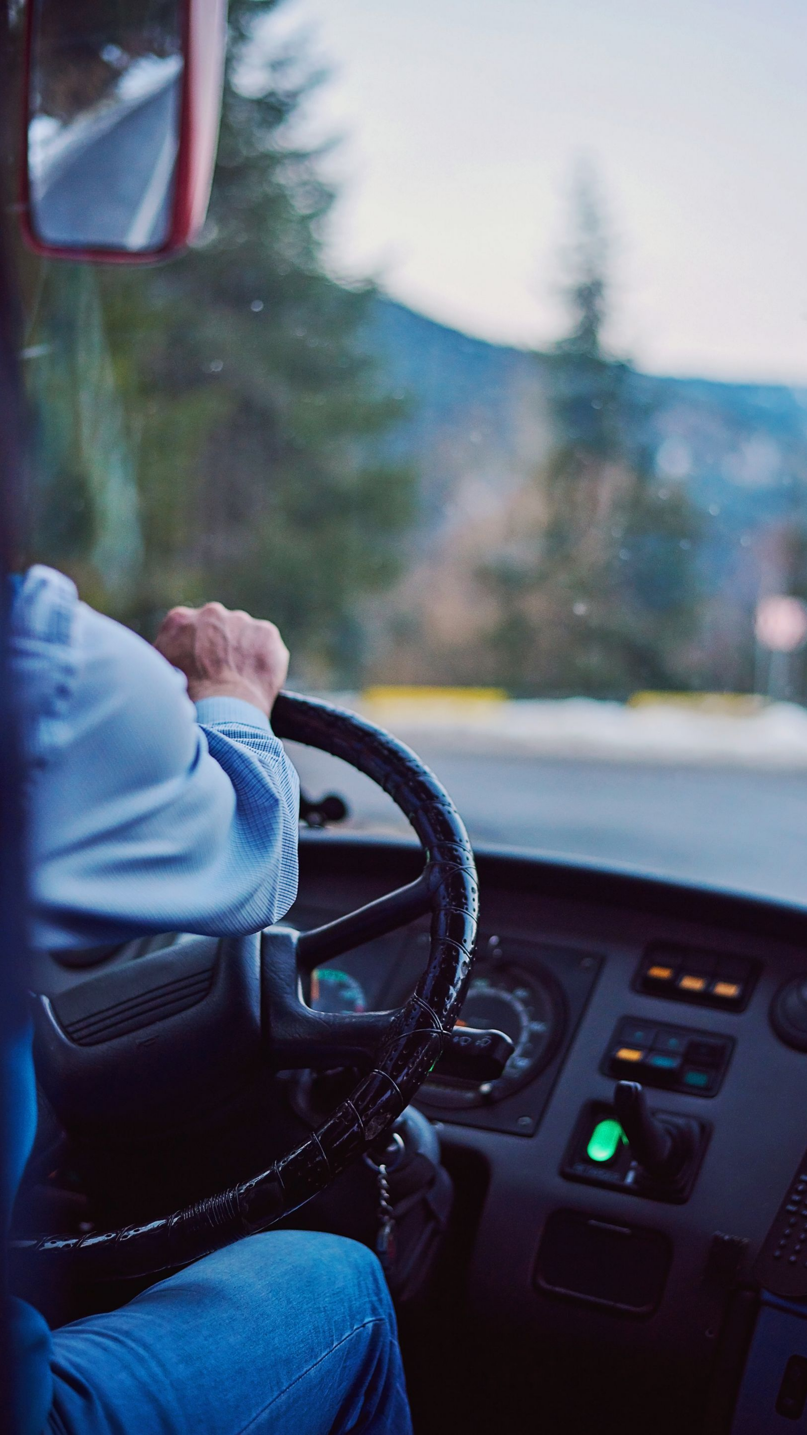 Une personne conduisant un bus, les mains sur le volant, la route et les montagnes visibles à travers le pare-brise.