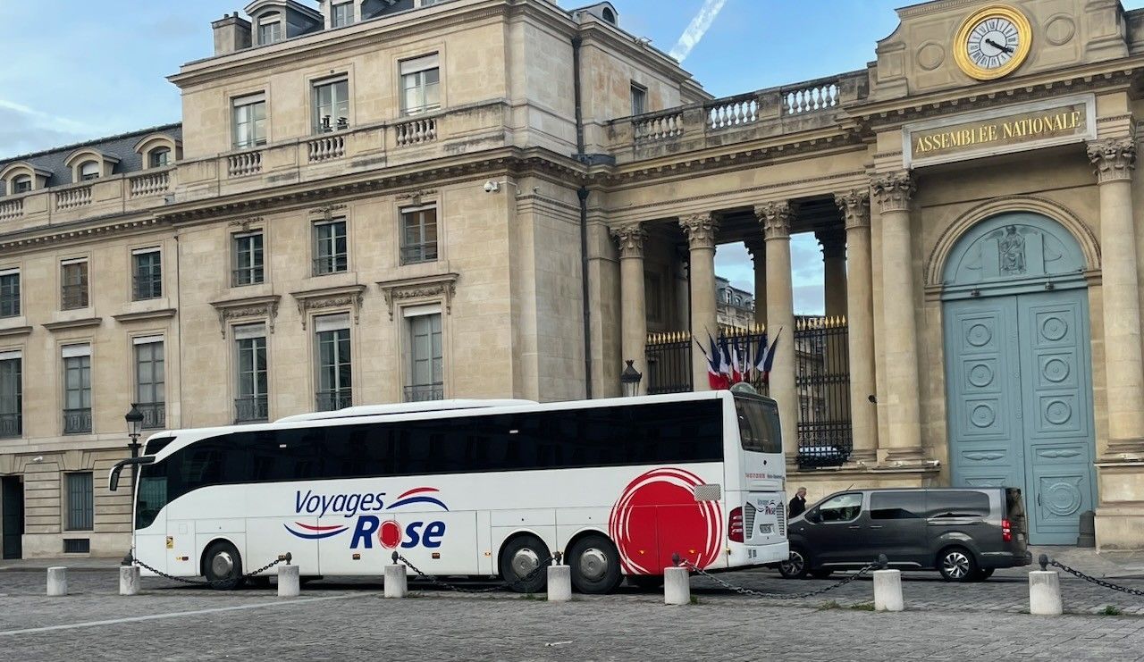 Un bus blanc des Voyages Rose est stationné devant l'entrée de l'Assemblée Nationale à Paris.