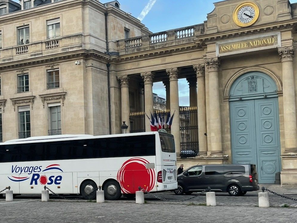 Un bus blanc des Voyages Rose est stationné devant l'entrée de l'Assemblée Nationale à Paris.