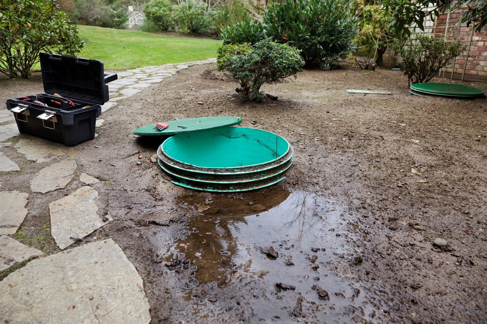 Cour inondée avec une couverture verte ronde, des flaques de boue, un chemin de pierres et une boîte à outils noire ouverte.