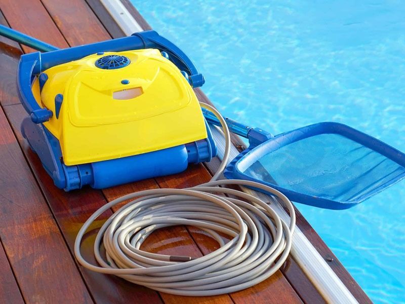 Una aspiradora robótica amarilla y azul está sentada en una terraza de madera al lado de una piscina.