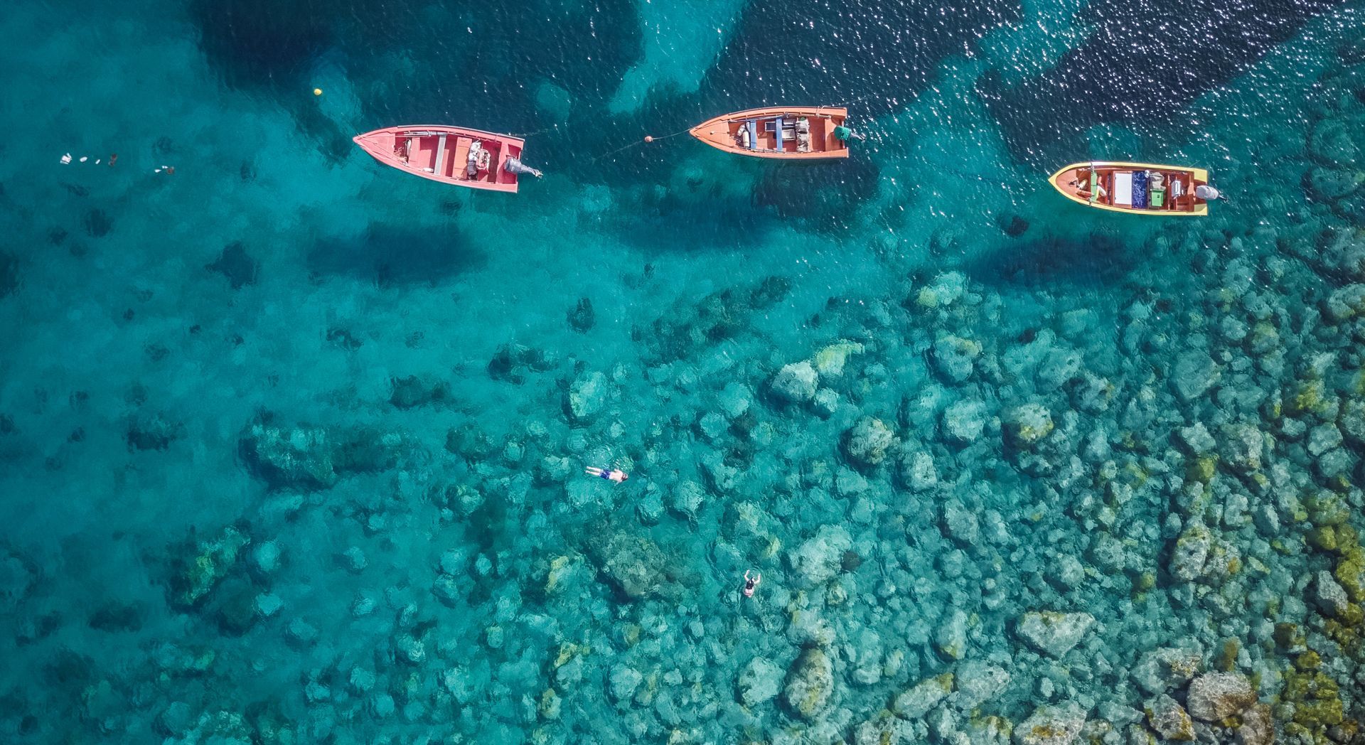 Trois bateaux colorés flottant sur une eau turquoise limpide, au-dessus d’un fond marin rocheux