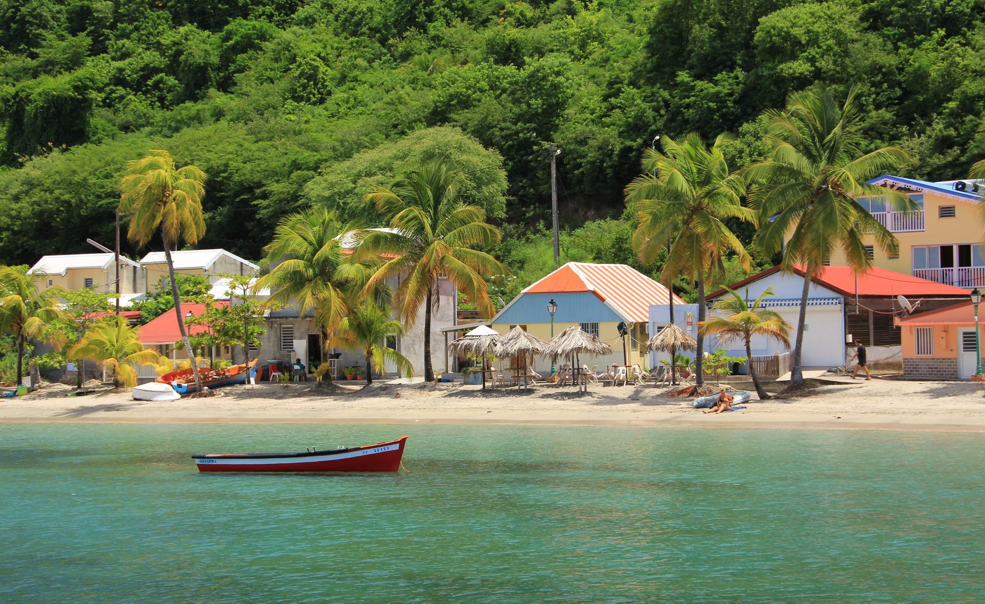 Plage tropicale avec des palmiers, de petites maisons colorées et un bateau rouge sur une eau turquoise limpide
