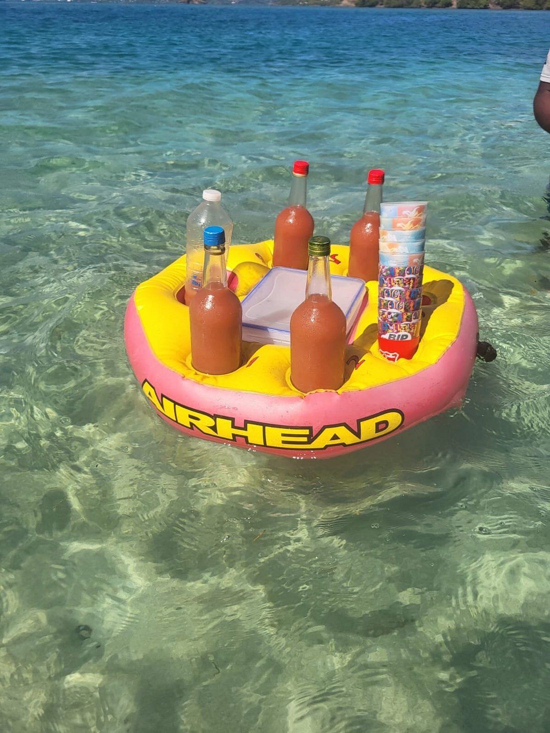 Radeau gonflable coloré avec des boissons, flottant sur une eau claire et peu profonde près d’une plage.