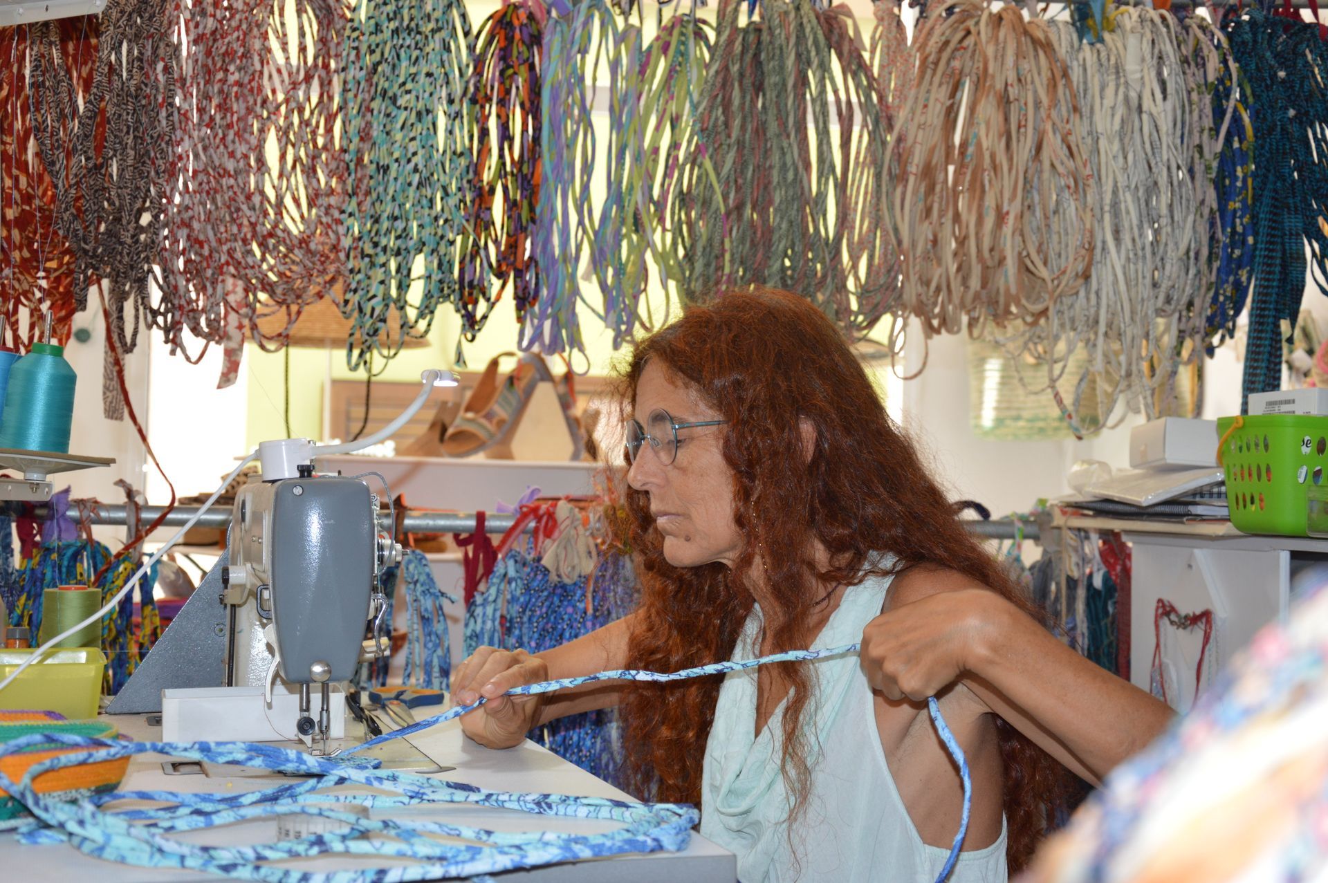 Una persona con cabello largo y rojizo utiliza una máquina de coser, rodeada de numerosos hilos de tela de colores que cuelgan del techo.