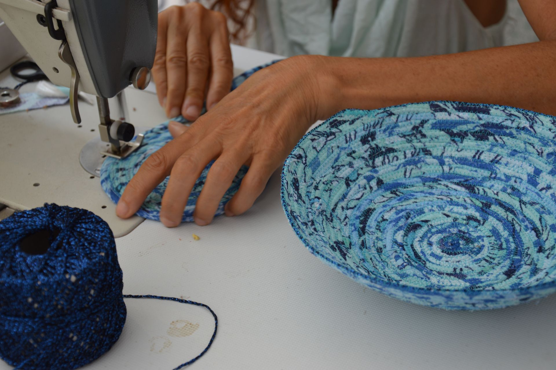 Una persona utiliza una máquina de coser para unir las piezas de un cuenco de tela azul enrollada. Un ovillo de hilo azul a juego se encuentra cerca.