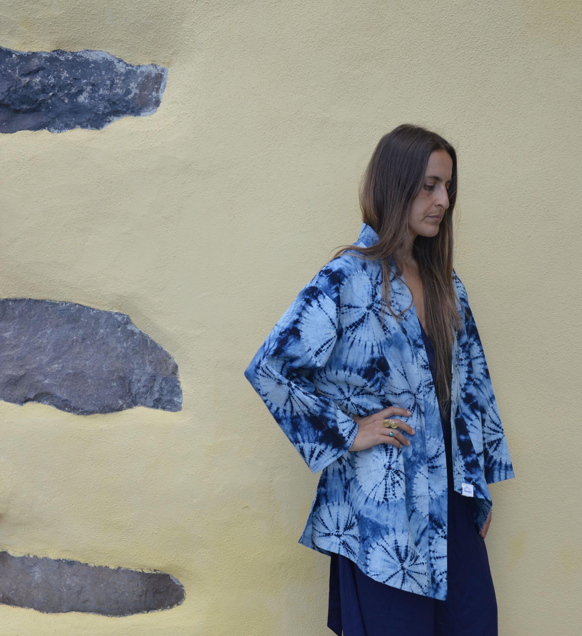 Mujer con chaqueta azul estampada posando contra una pared amarilla con detalles de piedra gris.