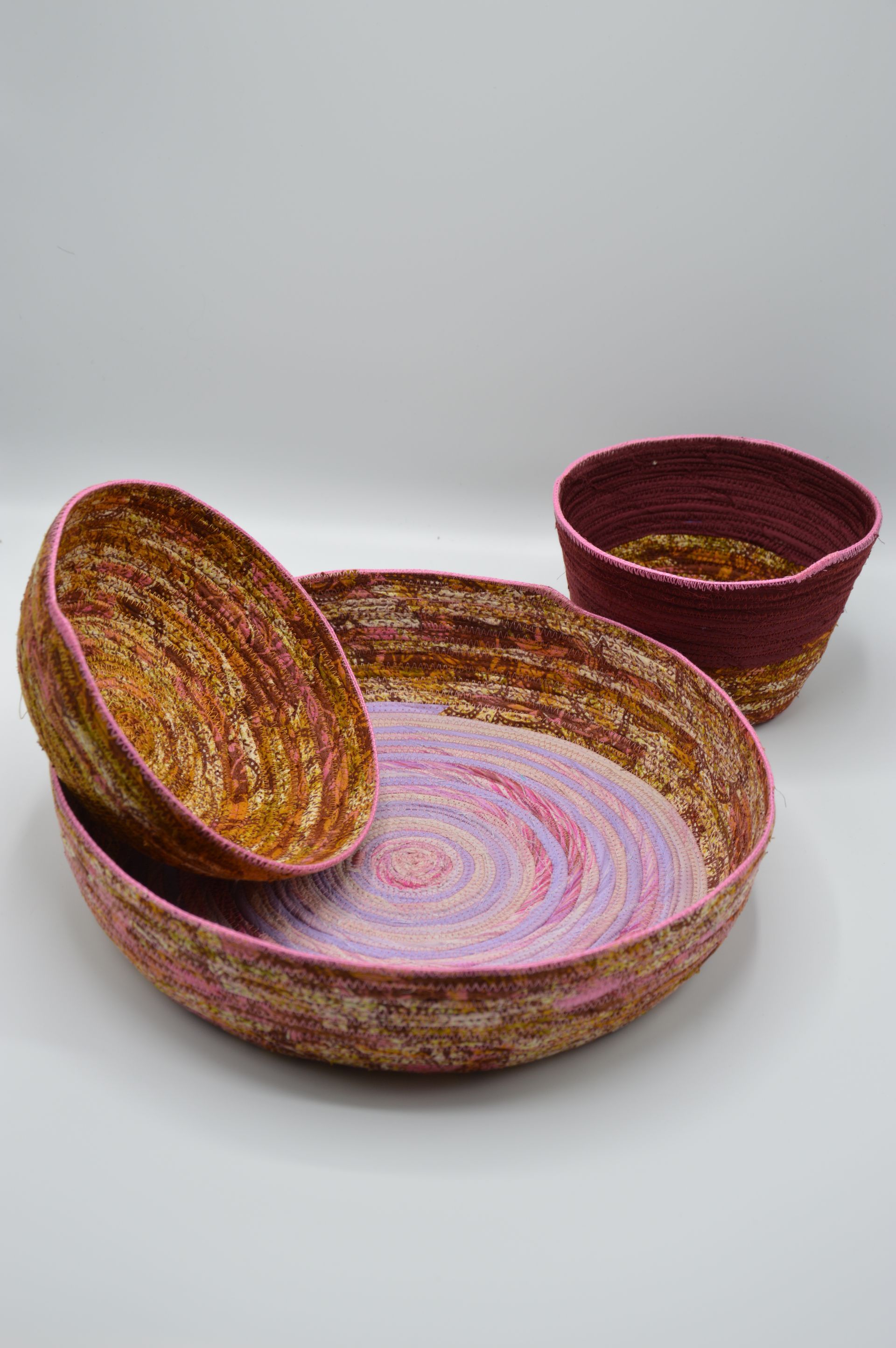 Tres cuencos de tela enrollada hechos a mano, de diferentes tamaños, con tonalidades de rosa, dorado y magenta sobre un fondo blanco.