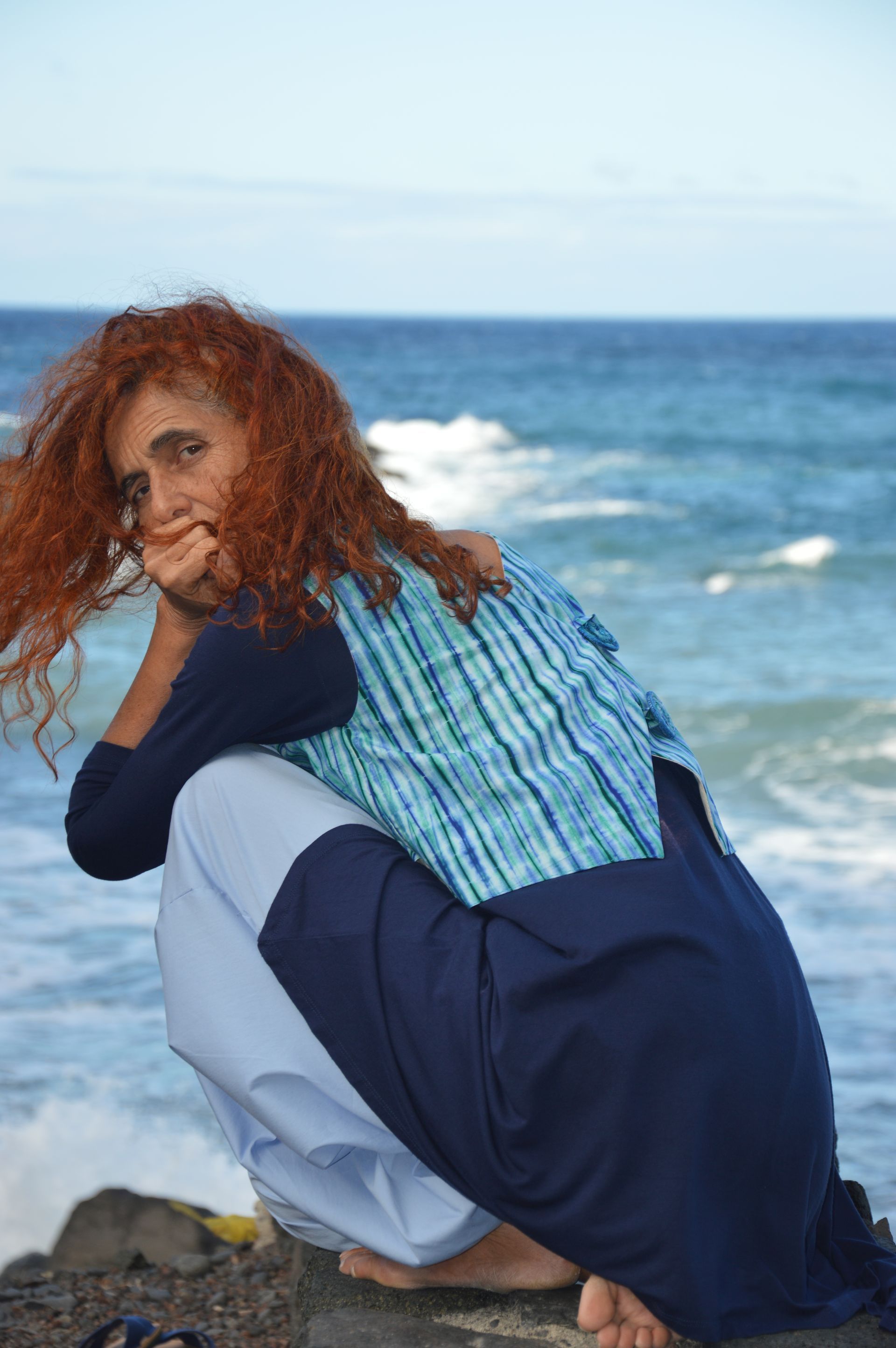 Persona agachada sobre unas rocas junto al mar, vestida con una blusa de rayas azules y una falda azul marino, con el pelo rizado y rojo.