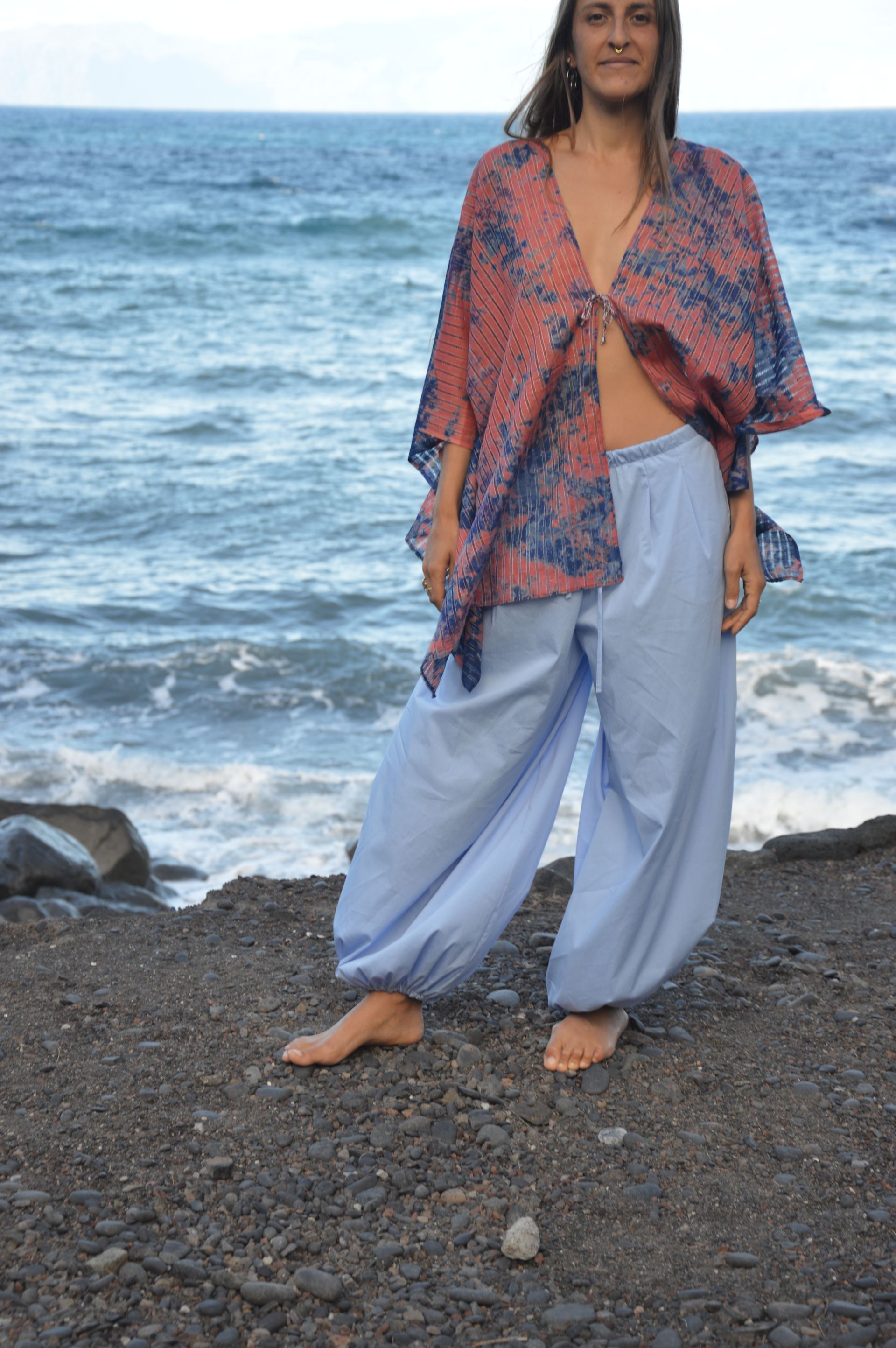 Mujer de pie en una playa rocosa, con una blusa estampada y pantalones azul claro, con las olas del océano de fondo.